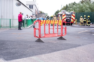 A red barrier stands in the foreground on an asphalt surface, with people gathered in the background near emergency vehicles. Several individuals are wearing uniforms, including high-visibility and firefighter gear. A firetruck with chevron markings is visible, and people are observing an activity. Green fencing and a metal building are also present in the scene.