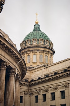 A large, ornate building with intricate architectural details and a green domed roof topped by a golden cross. The facade features tall columns and classical design elements, likely suggesting a religious or historical institution.