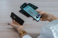 A person holds two smartphones, one in each hand, with attention focused on the device in their right hand. The smartphone screen displays a photo being taken of an object on a table. There are additional mobile phones visible on the table surface.