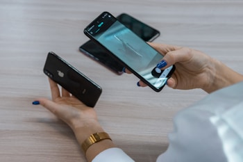A person holds two smartphones, one in each hand, with attention focused on the device in their right hand. The smartphone screen displays a photo being taken of an object on a table. There are additional mobile phones visible on the table surface.