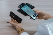 A person holds two smartphones, one in each hand, with attention focused on the device in their right hand. The smartphone screen displays a photo being taken of an object on a table. There are additional mobile phones visible on the table surface.