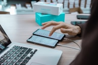 A desk with an open laptop and two smartphones side by side, both connected to charging cables. A hand is interacting with one of the smartphones. In the background, a box with 'Galaxy A32' printed on it is partially visible.