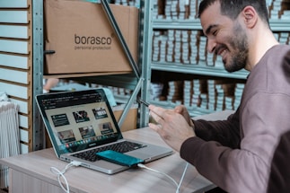 A person is seated at a desk in a workspace, smiling while interacting with their smartphone. A laptop is open in front of them, displaying a webpage. Shelves filled with boxes are visible in the background, and a large cardboard box labeled 'borasco personal protection' is placed next to the desk.