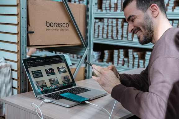 A person is seated at a desk in a workspace, smiling while interacting with their smartphone. A laptop is open in front of them, displaying a webpage. Shelves filled with boxes are visible in the background, and a large cardboard box labeled 'borasco personal protection' is placed next to the desk.