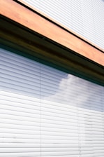A close-up view of a window with horizontal blinds partially closed, reflecting a faint image of clouds. The blinds are framed by a wooden border at the top.