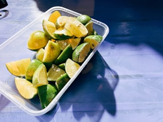 A rectangular plastic container filled with sliced lime wedges is placed on a blue surface. The limes have a vibrant green and yellow hue, with some of the sections showing the juicy pulp. Shadows cast by the container and limes create an interesting contrast on the surface.