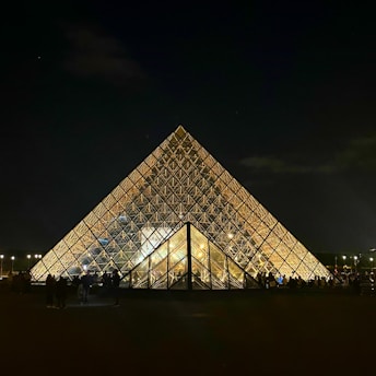 A large glass pyramid structure is illuminated against a dark night sky. Its geometric panels reflect the light, creating a golden glow. People are gathered around, silhouetted against the bright pyramid. The atmosphere is both grand and tranquil.