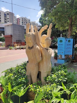 A wooden sculpture of three figures with raised arms is situated in a landscaped area with green plants. The setting is urban, with high-rise buildings in the background and a blue sky overhead. Nearby, there are informational boards from the New Delhi Municipal Council.
