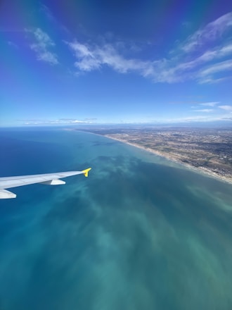 Aerial view of a coastline with a clear, expansive blue sky and scattered clouds. The vibrant turquoise sea stretches along the shore, and part of an airplane wing with a yellow tip is visible. The land features a mix of urban and natural areas.