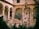 An elegant courtyard with arches supported by ornate columns. In the center, there is a large stone well with a metal frame and pulley system. Several people are gathered near the well, bordered by lush green trees, creating a serene and historical ambiance.