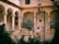 An elegant courtyard with arches supported by ornate columns. In the center, there is a large stone well with a metal frame and pulley system. Several people are gathered near the well, bordered by lush green trees, creating a serene and historical ambiance.