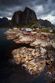 A dramatic landscape featuring a cluster of traditional Nordic red houses nestled along rugged, moss-covered rocky cliffs by the sea. A towering mountain looms in the background under a moody sky filled with dark, swirling clouds.