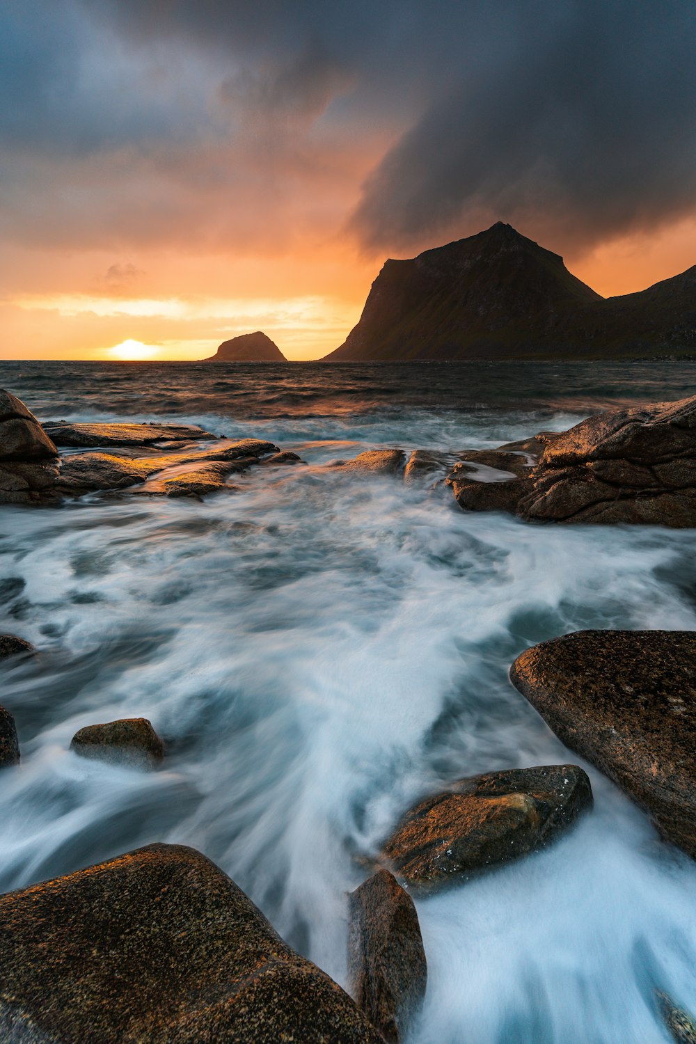 the sun is setting over the ocean with rocks in the foreground