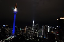 A nighttime cityscape showcasing a skyline illuminated by city lights, featuring a prominent telecommunications tower glowing with blue and white lights. The surrounding skyscrapers, including a tapered building with bright lights, enhance the urban atmosphere.
