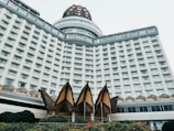 A large, multi-story hotel building with a modern architectural design. The structure features a white facade with numerous windows arranged in a symmetrical pattern. Near the base, triangular roof structures and lush greenery are visible.