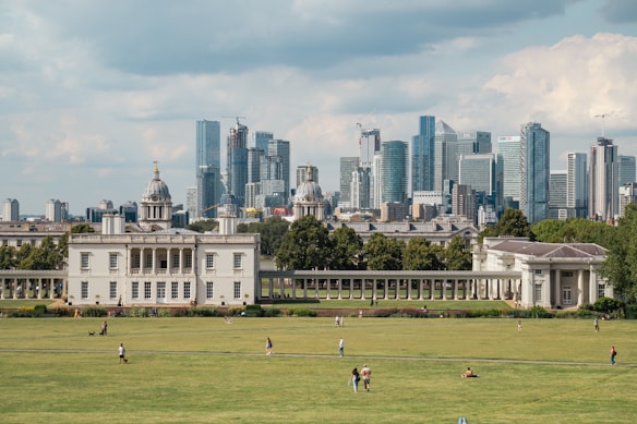 In the foreground, a historic classical building is surrounded by expansive green lawns with a few people walking and sitting. Beyond this, a cityscape of modern skyscrapers dominates the background, featuring glass and metal structures.