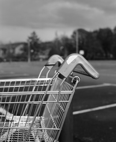 A close-up of a shopping cart with a blurred background of a parking lot and trees. The cart's handle is visible, emphasizing its metal structure.