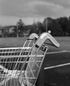 A close-up of a shopping cart with a blurred background of a parking lot and trees. The cart's handle is visible, emphasizing its metal structure.