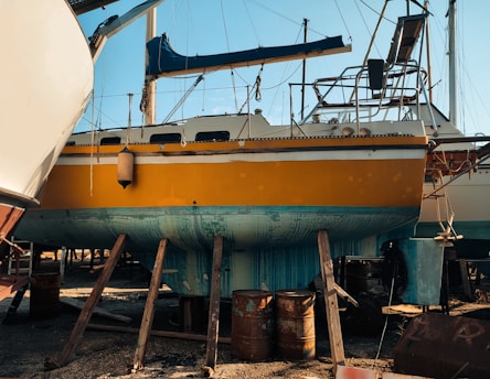 A bright yellow boat is elevated on wooden supports in a shipyard, with parts of its exterior showing wear and patches of teal paint. Various equipment and barrels are scattered around, and other boats are visible in the background against a clear blue sky.