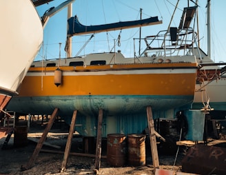 A bright yellow boat is elevated on wooden supports in a shipyard, with parts of its exterior showing wear and patches of teal paint. Various equipment and barrels are scattered around, and other boats are visible in the background against a clear blue sky.