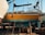 A bright yellow boat is elevated on wooden supports in a shipyard, with parts of its exterior showing wear and patches of teal paint. Various equipment and barrels are scattered around, and other boats are visible in the background against a clear blue sky.