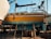 A bright yellow boat is elevated on wooden supports in a shipyard, with parts of its exterior showing wear and patches of teal paint. Various equipment and barrels are scattered around, and other boats are visible in the background against a clear blue sky.
