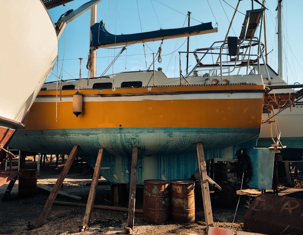 A bright yellow boat is elevated on wooden supports in a shipyard, with parts of its exterior showing wear and patches of teal paint. Various equipment and barrels are scattered around, and other boats are visible in the background against a clear blue sky.