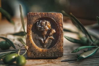 A rustic rectangular plaque with an embossed design of a flower, situated on a wooden surface. The plaque has a worn, antique appearance with earthy tones and the text 'To your health' engraved at the bottom. Olive branches and green olives are arranged around the plaque, contributing to a natural and organic setting.