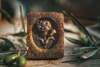 A rustic rectangular plaque with an embossed design of a flower, situated on a wooden surface. The plaque has a worn, antique appearance with earthy tones and the text 'To your health' engraved at the bottom. Olive branches and green olives are arranged around the plaque, contributing to a natural and organic setting.