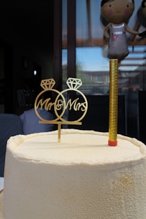 A textured white cake with a gold decorative topper reading 'Mr & Mrs' next to a single gold candle. Behind the cake, a large window shows reflections and part of a balloon resembling a person.