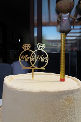 A textured white cake with a gold decorative topper reading 'Mr & Mrs' next to a single gold candle. Behind the cake, a large window shows reflections and part of a balloon resembling a person.