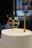 A textured white cake with a gold decorative topper reading 'Mr & Mrs' next to a single gold candle. Behind the cake, a large window shows reflections and part of a balloon resembling a person.