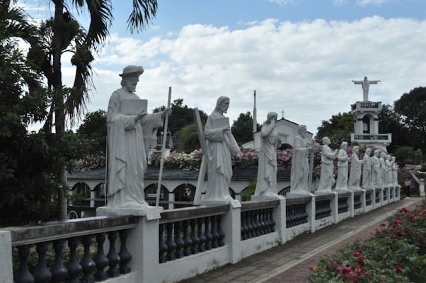 A row of white statues depicting religious figures, possibly saints or apostles, lined up along a stone balustrade in an outdoor setting. The background reveals lush greenery and flowering plants, with a prominent larger statue of a figure on a pedestal visible in the distance.