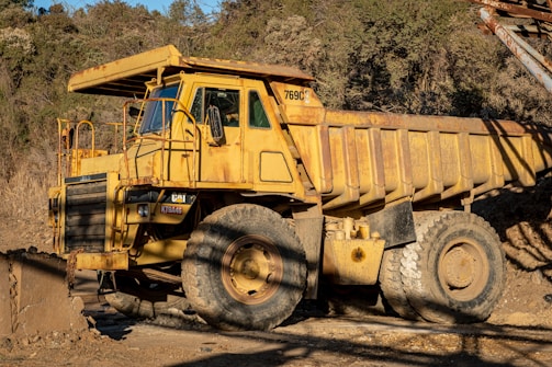 An industrial dump truck with a yellow, weathered exterior parked on rugged terrain near a forested area. The massive vehicle features large, thick tires and is designed for heavy-duty tasks.