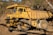 An industrial dump truck with a yellow, weathered exterior parked on rugged terrain near a forested area. The massive vehicle features large, thick tires and is designed for heavy-duty tasks.