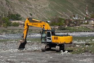 A yellow Hyundai excavator with a large bucket is parked on rocky terrain next to a small river. The background features a hilly landscape with sparse vegetation and a few houses and structures. There's a telecommunications tower visible on the right side.