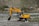 A yellow Hyundai excavator with a large bucket is parked on rocky terrain next to a small river. The background features a hilly landscape with sparse vegetation and a few houses and structures. There's a telecommunications tower visible on the right side.