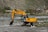 A yellow Hyundai excavator with a large bucket is parked on rocky terrain next to a small river. The background features a hilly landscape with sparse vegetation and a few houses and structures. There's a telecommunications tower visible on the right side.