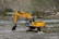 A yellow Hyundai excavator with a large bucket is parked on rocky terrain next to a small river. The background features a hilly landscape with sparse vegetation and a few houses and structures. There's a telecommunications tower visible on the right side.