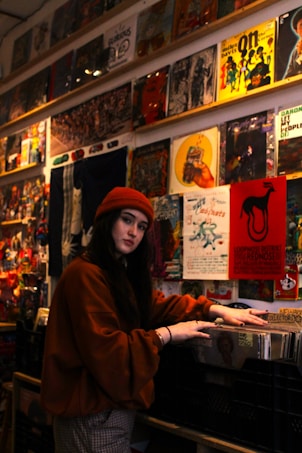 A person wearing a red beanie and dark orange sweater stands in a shop or store surrounded by a collection of vintage vinyl records on display. The walls are adorned with colorful retro posters and album covers.