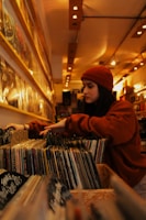 A person wearing a red beanie and a matching sweatshirt browses through vinyl records in a cozy, warmly lit record store. The records are neatly organized on wooden shelves that stretch along the length of the wall. Overhead lights create a comfortable ambiance, highlighting the variety of album covers.