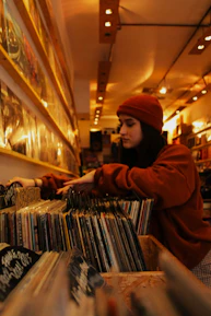 A person wearing a red beanie and a matching sweatshirt browses through vinyl records in a cozy, warmly lit record store. The records are neatly organized on wooden shelves that stretch along the length of the wall. Overhead lights create a comfortable ambiance, highlighting the variety of album covers.