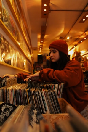 A person wearing a red beanie and a matching sweatshirt browses through vinyl records in a cozy, warmly lit record store. The records are neatly organized on wooden shelves that stretch along the length of the wall. Overhead lights create a comfortable ambiance, highlighting the variety of album covers.