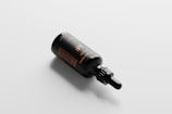 A dark brown or black dropper bottle lies on its side on a light gray, flat surface. The bottle has a glossy finish with gold text printed on it, which indicates some product-related information.