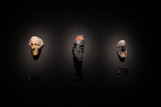 Three masks are displayed against a dark background. The mask on the left has a pale, human-like face with an elongated chin. The center mask is darker with a gas mask-like appearance and orange stripe along the top. The right mask is stone-like, anthropomorphic with rough textures.