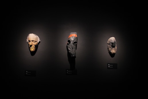 Three masks are displayed against a dark background. The mask on the left has a pale, human-like face with an elongated chin. The center mask is darker with a gas mask-like appearance and orange stripe along the top. The right mask is stone-like, anthropomorphic with rough textures.