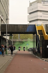 A bustling urban scene with people walking along a pathway. Motorcyclists are riding forward, and bicycles are parked on the side. Green smoke drifts across the road as tall buildings and an elevated walkway frame the view.