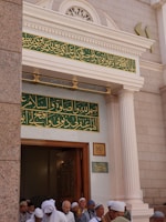 A group of people wearing traditional attire gather at the entrance of a building with intricate Arabic calligraphy above the doorway. The architectural style features ornate columns and decorative elements around the entrance.