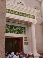 A group of people wearing traditional attire gather at the entrance of a building with intricate Arabic calligraphy above the doorway. The architectural style features ornate columns and decorative elements around the entrance.