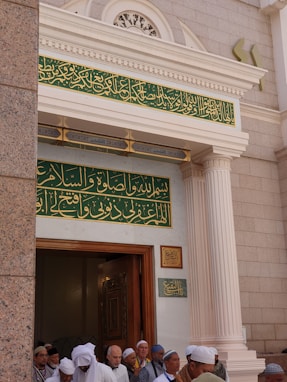 A group of people wearing traditional attire gather at the entrance of a building with intricate Arabic calligraphy above the doorway. The architectural style features ornate columns and decorative elements around the entrance.
